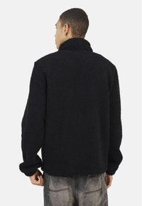 Giacca in pile nera con una finitura testurizzata e morbida, dotata di un colletto alto e una vestibilità comoda. Maniche lunghe con dettagli ai polsini, indossata con jeans grigi.