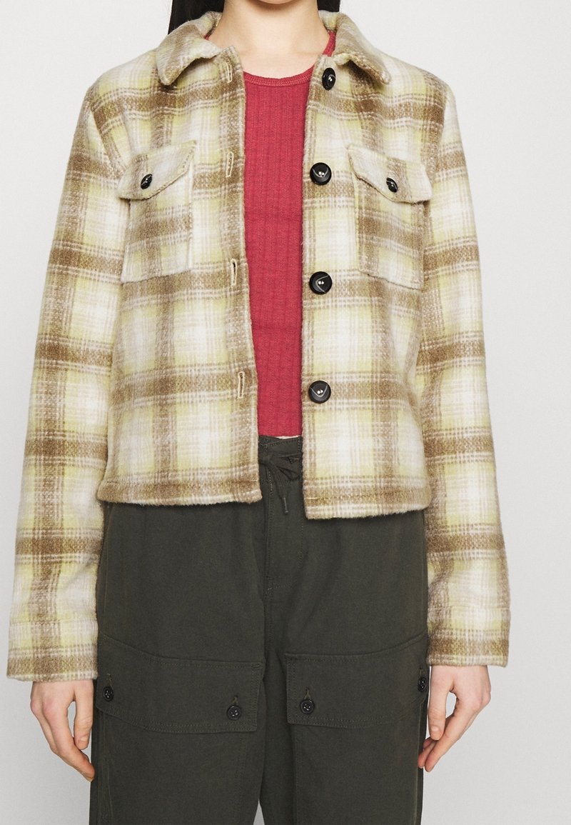 Fleecefodrad jacka i beige och grön rutigt, med krage, knappar framtill, två bröstfickor, kombinerad med en burgunderröd topp och mörka byxor.