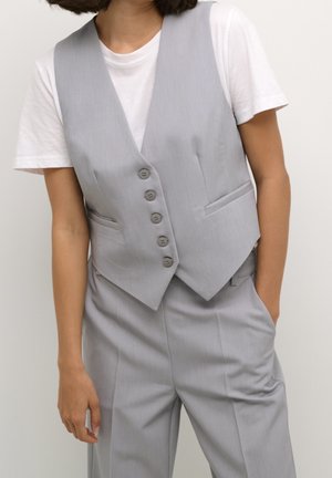 Personne portant une veste gris clair boutonnée par-dessus un t-shirt blanc à manches courtes avec un pantalon gris assorti, une main dans la poche.