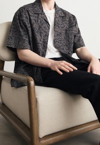 Korte mouw shirt in donkergrijs met een subtiel abstract patroon, gecombineerd met een wit t-shirt en zwarte broek, zittend op een lichtgekleurde fauteuil.