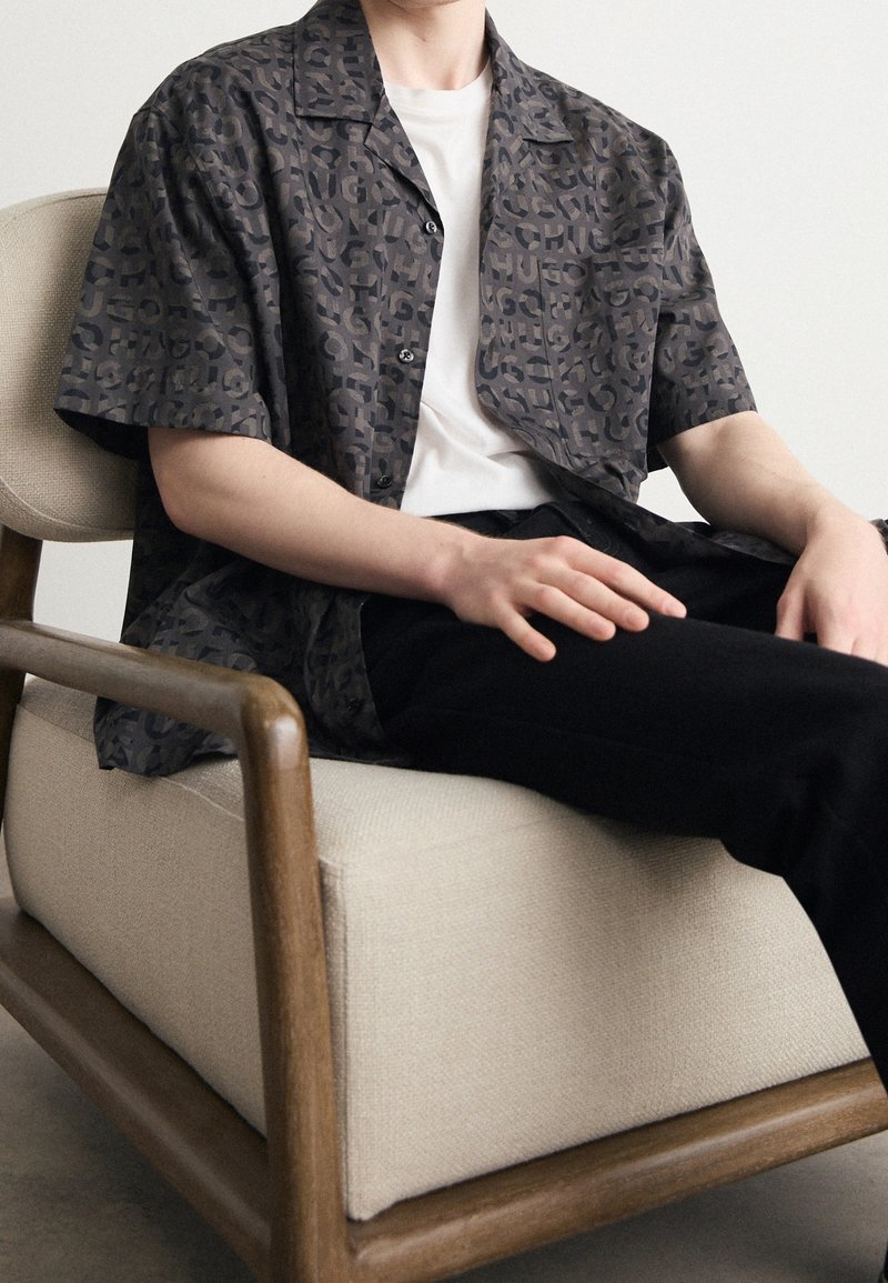 Korte mouw shirt in donkergrijs met een subtiel abstract patroon, gecombineerd met een wit t-shirt en zwarte broek, zittend op een lichtgekleurde fauteuil.