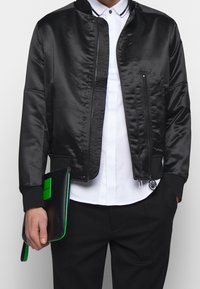 Svart satin bomberjacka med dragkedja, ribbade ärmslut och snedställda sidofickor, bärs över en vit skjorta och hålls med en clutch med gröna detaljer.