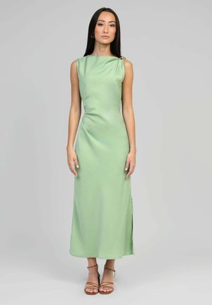 Femme aux longs cheveux foncés portant une robe sans manches en satin vert clair et des talons bruns à lanières, debout devant un fond uni gris clair.