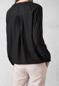Schwarze, strukturierte Bluse mit langen Ärmeln, gerafftem Rückendetail und geknöpften Manschetten, kombiniert mit hellen, taillierten Hosen.