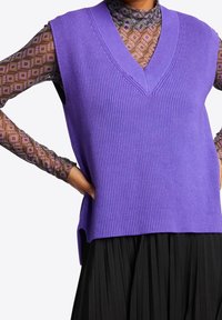 Lila gerippter, ärmelloser Pullover mit V-Ausschnitt, getragen über einem gemusterten Langarmoberteil, kombiniert mit einem schwarzen plissierten Rock.