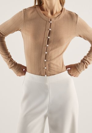 Woman wearing a beige ribbed cardigan with pearl buttons paired with high-waisted white pants, hands resting on hips.