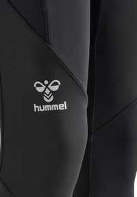 Svart våtdräkt med slät yta, utrustad med ett nätpanel och flexibla sömmar. På dräkten visas det vita "hummel"-logotypen och designaccenter.