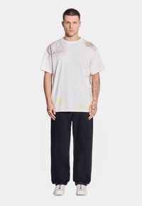 Biała koszulka z grafiką w abstrakcyjne wzory w odcieniach różu i żółci, połączona z ciemnymi dżinsami i białymi sneakersami.