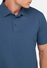 Zils polo krekls, kas izgatavots no mīkstas, struktūrētas auduma. Iezīmē klasisku apkakli, trīs pogu aizdari un īsās piedurknes, nodrošinot pieguļošu izskatu.
