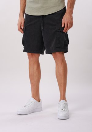 Man wearing black corduroy cargo shorts and white sneakers, standing against a plain light background.