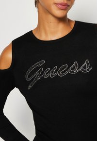 Zwarte geribbelde lange mouwen top met een off-shoulder ontwerp en "Guess" versierd met rhinestones in cursieve letters.