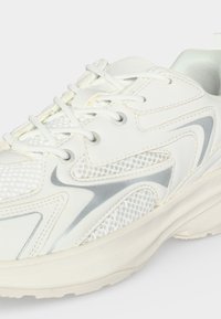 Baskets de sport blanches avec panneaux en maille, accents en forme de vague argentés, lacets blancs et semelle blanche épaisse.