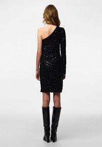 Czarna mini sukienka na jedno ramię z teksturowanym wzorem z cekinów; z długim rękawem po jednej stronie. Modelka nosi czarne, wysokie botki na obcasie sięgające kolan.