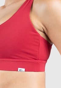 Sujetador deportivo rojo hecho de tela suave con un escote profundo y una banda inferior ancha. Presenta una pequeña etiqueta de la marca en la parte inferior izquierda.