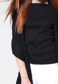 Svart figursydd topp i kombination med en svävande svart cardigan. Toppen har en rund halsringning och samlad textur, som framhävs mot vita byxor.