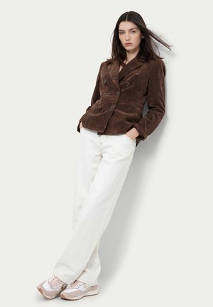 Donna con capelli scuri che indossa una giacca doppiopetto marrone, pantaloni bianchi a gamba larga e scarpe da ginnastica rosa e bianche, in piedi con le mani nelle tasche.