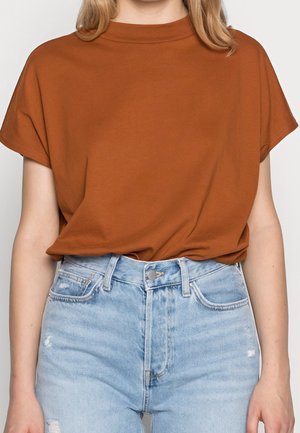 Kvinde iført en rustfarvet kortærmet skjorte, stoppet ned i lyseblå, slidte, højtaljede jeans mod en ensfarvet baggrund.