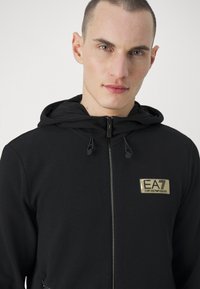 Giacca nera con zip e finitura testurizzata, dotata di cappuccio. Presenta un patch con il logo dorato EA7 sul petto e cordini sul cappuccio per la regolazione.