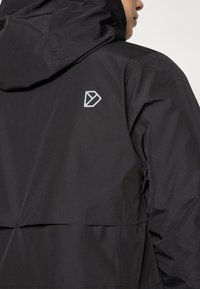 Čierna nepremokavá bunda s kapucňou, na zadnej strane s strieborným geometrickým logom, hladká textúra a aerodynamický dizajn.