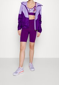 Veste de sport violette avec des accents violets clairs, soutien-gorge de sport floral, short de cyclisme violet assorti et baskets violettes claires avec des détails orange.