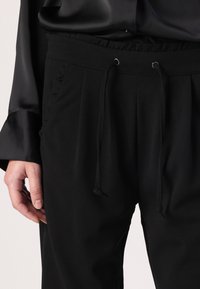 Svarta byxor med lös passform, med elastisk midja, dragsko och sidofickor. Tyget ser slätt ut med en matt yta.