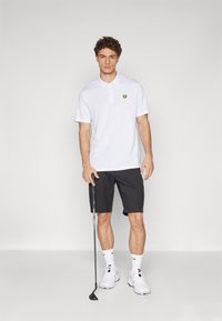 White polo shirt with a yellow logo, paired with black shorts. Model holds a golf club, wearing white shoes and socks.