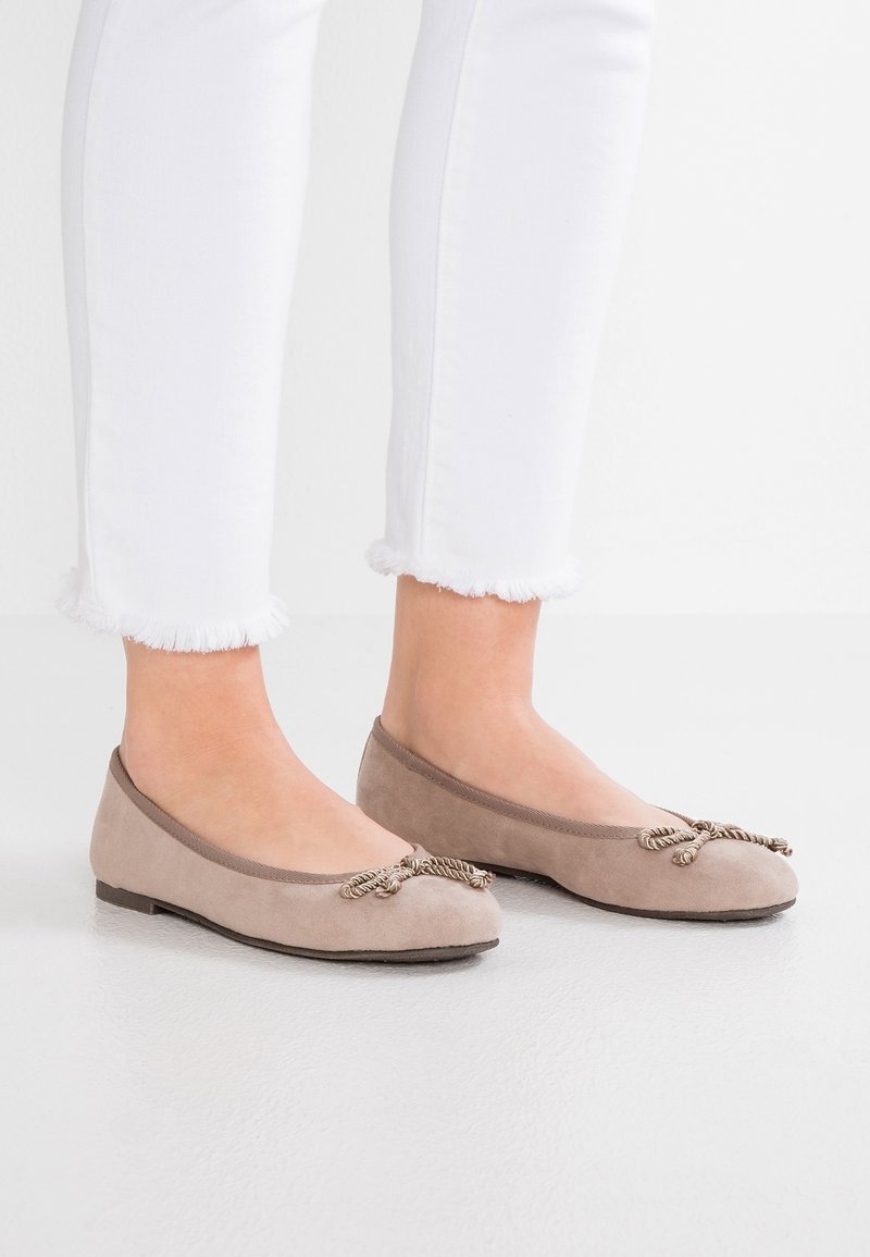 Jambes portant un pantalon blanc à ourlet effiloché et des ballerines en daim beige avec des nœuds décoratifs en corde torsadée sur un sol clair.
