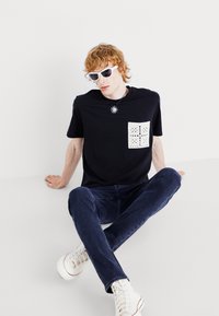 T-shirt noir avec une poche en dentelle blanche, design ajusté, associé à un jean foncé et des baskets montantes blanches, porté avec des lunettes de soleil et un collier.