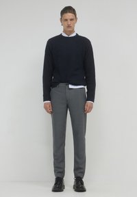 Maglione a maglia blu navy, con texture a coste; camicia bianca con colletto sotto. Pantaloni grigi a fantasia, slim fit. Scarpe nere con lacci. Sfondo semplice.