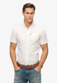 Camicia bianca a maniche corte con colletto a coreano, dotata di tasca sul petto. Abbinata a jeans blu scoloriti e una cintura in pelle marrone.