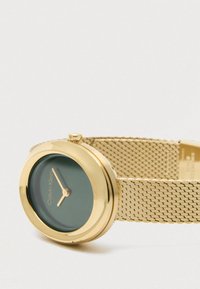 Złoty zegarek Calvin Klein na nadgarstek z czarną okrągłą tarczą i złotą siateczkową bransoletą, wskazujący godzinę 10:10.