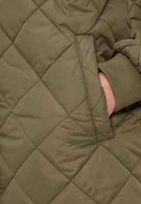 Πράσινο χακί τζάκετ με ραμμένο σχέδιο σε σχήμα ρόμβου, με πλαϊνή τσέπη και λεπτομέρεια με ριμπ μανσέτα. Μαλακό, γυαλιστερό ύφασμα με μόνωση.