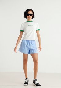 T-shirt bianca con collo verde e grafica, abbinata a pantaloni corti blu chiaro. Sneakers in bianco e nero completano l'outfit. Tessuti morbidi al tatto.