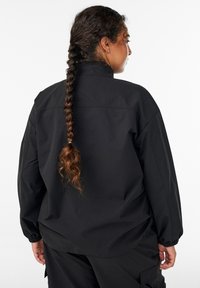 Musta ylisuurikokoinen takki, jossa on korkea kaulus, joustavat hihansuut ja sileä pinta. Takana näkyy letitetty kampaus.