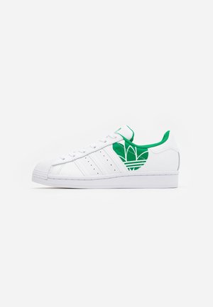 Chaussure de sport blanche avec des accents verts. Comprend un embout texturé, trois bandes latérales et une languette verte avec logo. Semelle plate en caoutchouc.