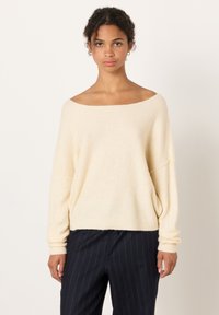 Kvinna klädd i en krämfärgad off-shoulder tröja och mörka byxor med kritstrecksrandar, stående mot en enkel ljus bakgrund.