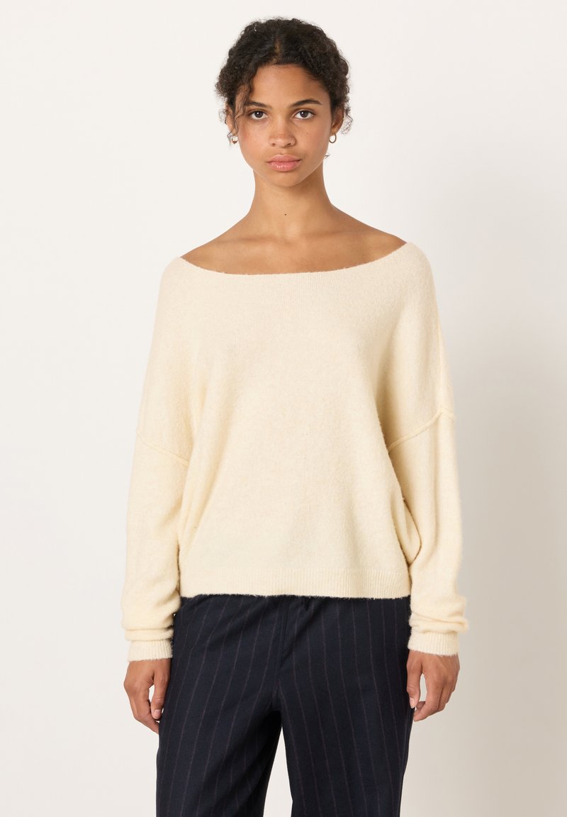 Kvinna klädd i en krämfärgad off-shoulder tröja och mörka byxor med kritstrecksrandar, stående mot en enkel ljus bakgrund.