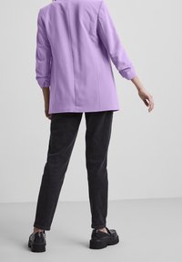 En person står vänd bort, iklädd en lavendelfärgad blazer, svarta byxor och svarta skor med kraftig sula, mot en enkel grå bakgrund.