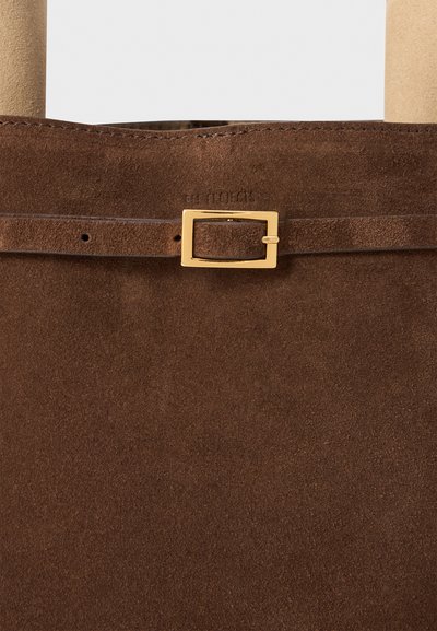 Sac en daim marron avec une surface texturée, mettant en avant une petite boucle en or et des détails de sangle. Bords cousus et marque embossée visibles.