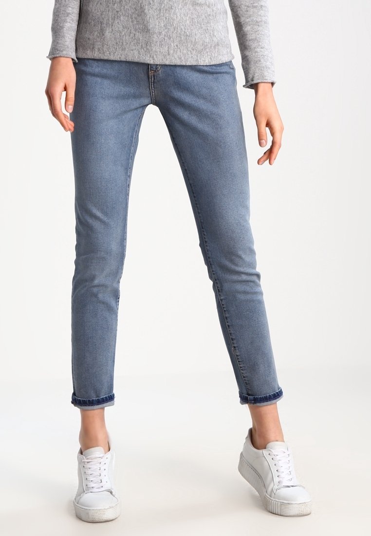 Jeans skinny de mezclilla azul claro con un ajuste clásico, puños enrollados y un ligero estiramiento. Llevados con zapatillas blancas sobre un fondo neutro.