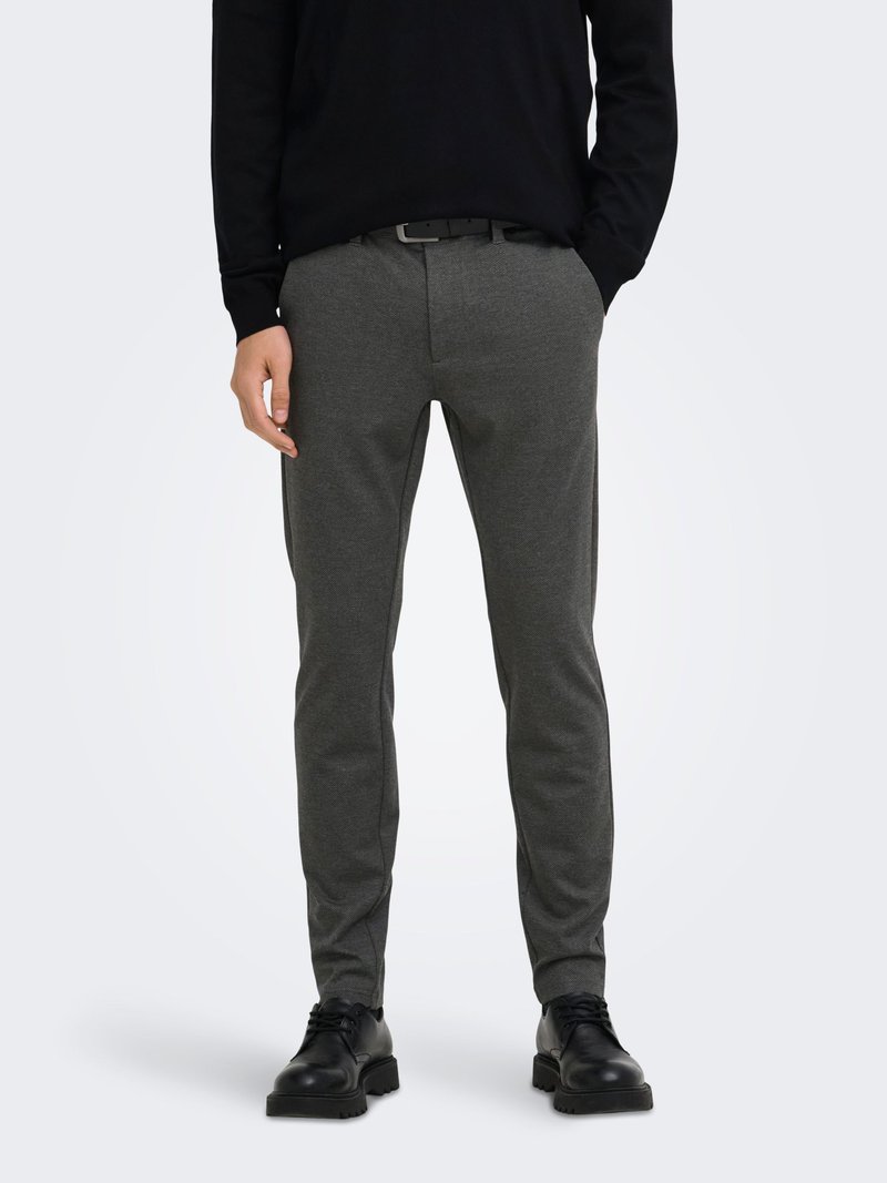 Homme portant un pantalon gris ajusté avec une ceinture noire, un pull noir et des chaussures de cuir noires à lacets, debout les mains dans les poches.