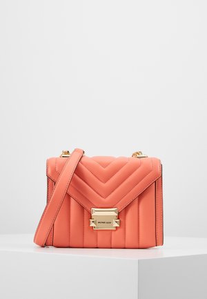 Sac bandoulière en cuir matelassé rose corail avec fermoir doré et détail chaîne, exposé sur un socle blanc sur fond uni.