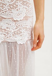 Main féminine reposant à côté d'une robe blanche en dentelle avec broderies florales et tissu transparent à pois.