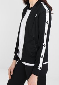 Black zip-up jacket with white contrasting side stripes featuring arrow patterns, textured fabric, and ribbed cuffs and collar.