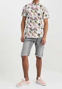 Hombre que lleva una camisa blanca de manga corta con formas abstractas coloridas, pantalones cortos grises arremangados y zapatillas rosas claras con rayas blancas.