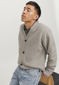 Cardigan lavorato a maglia di un grigio chiaro con scollo a V, tre bottoni scuri e polsini a costine; indossato sopra una camicia a scacchi abbinata a jeans blu.