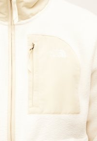 Μπουφάν από κρεμ φλις με φερμουάρ στην τσέπη του στήθους που φέρει το λογότυπο The North Face κεντημένο με λευκό χρώμα.