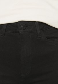 Svarta denimjeans med en mjuk textur, försedd med knappstängning fram och femficksdesign, delvis täckt av en ljusfärgad skjorta.