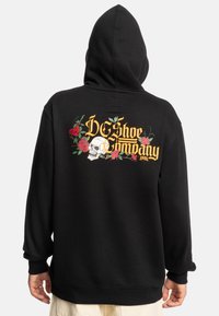 Sudadera negra con un gráfico de calavera y diseños florales en la parte posterior. Presenta puños y dobladillo acanalados, textura suave y un bolsillo tipo canguro en la parte frontal.