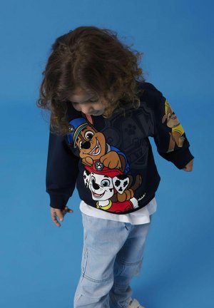 Felpa blu scuro con personaggi di cani dei cartoni animati; il tessuto è di cotone con grafiche stampate; abbinata a pantaloni in denim chiaro e una maglietta bianca sotto.